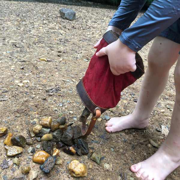 little boy standing on a rocky beach, dumping rocks out of a red waxed canvas and brown leather bucket bag