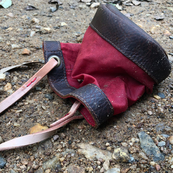 red waxed canvas and brown leather bucket bag laying on a rocky beach