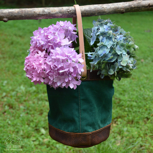 pink and blue hydrangeas in a green waxed canvas and brown leather bucket bag, hanging from a branch