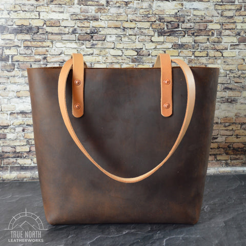 hand stitched brown leather tote on a dark surface with a faux brick background behind it